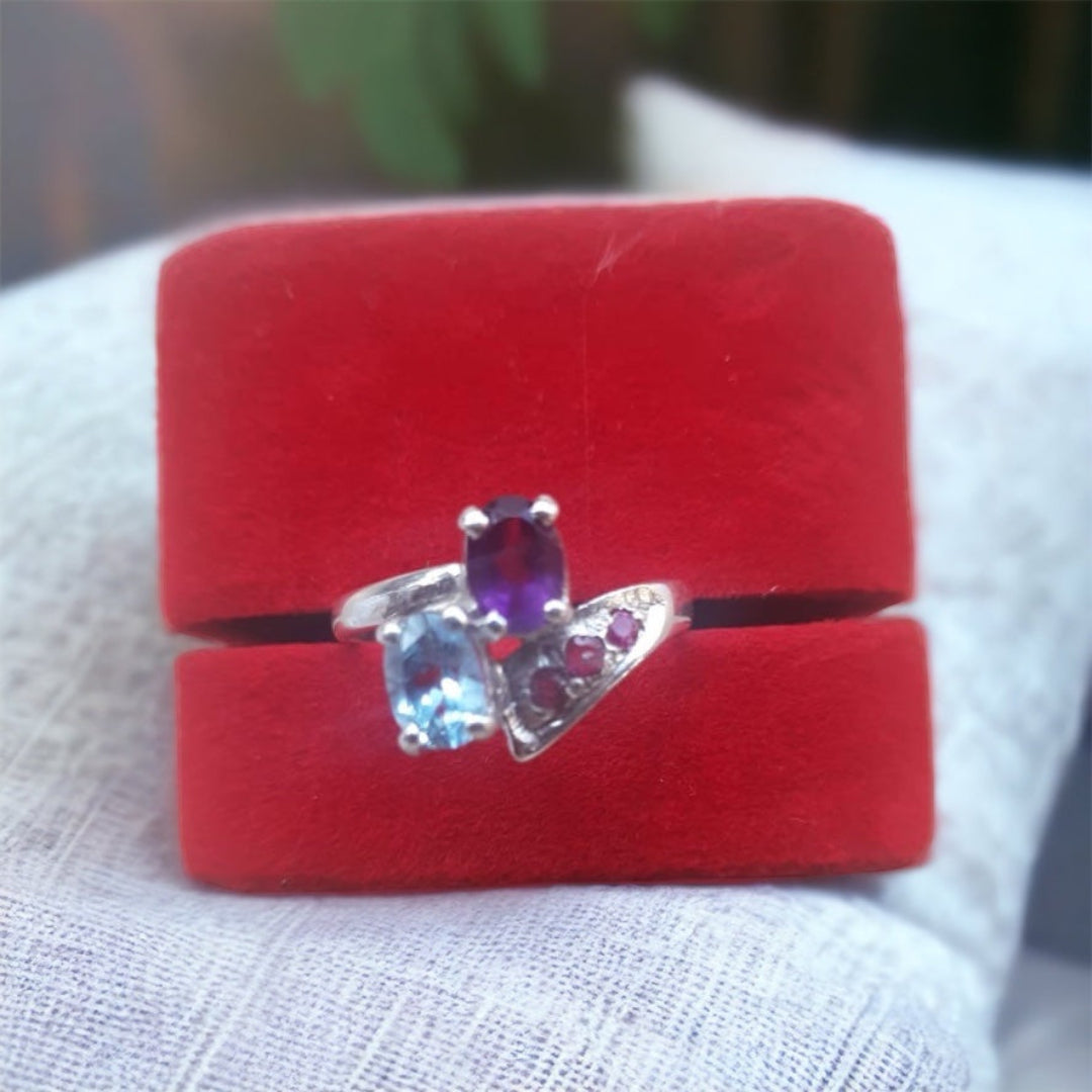 A silver ring featuring a blue topaz, amethyst, and pink sapphire set on a red cushion.